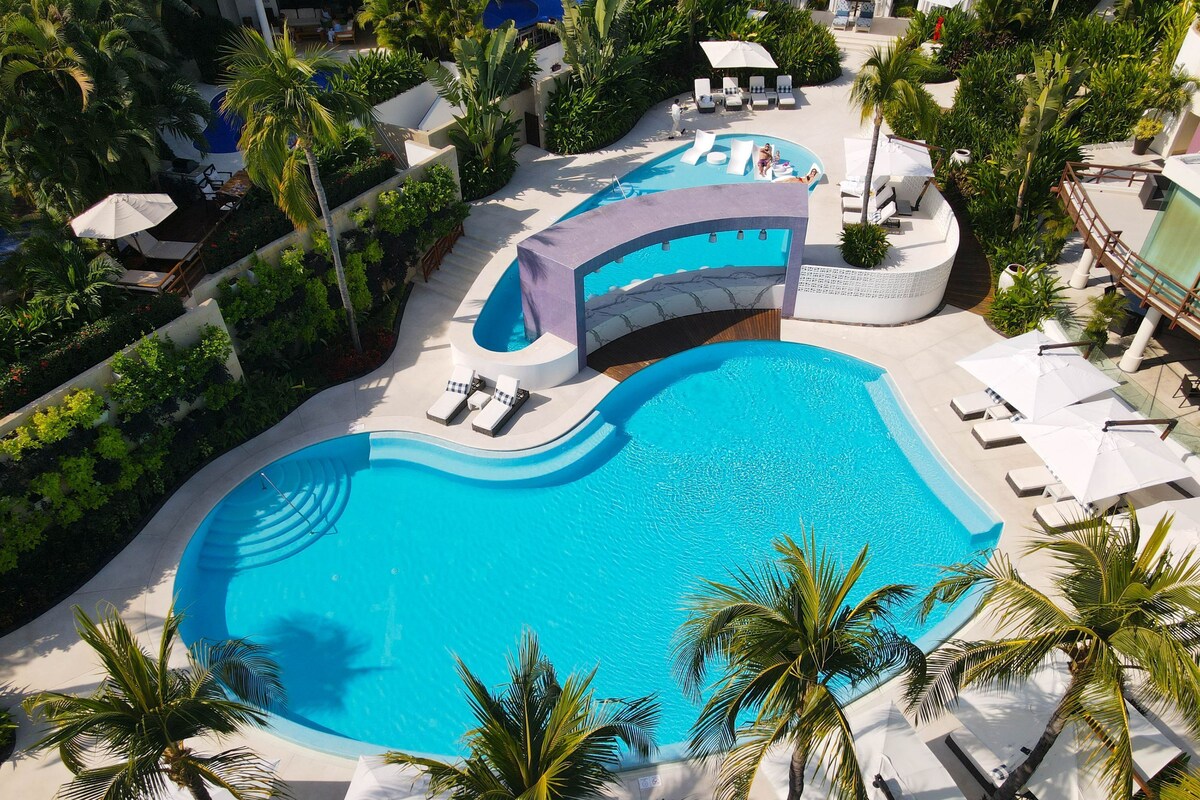 A resort with a large swimming pool, lounge chairs, and palm trees.