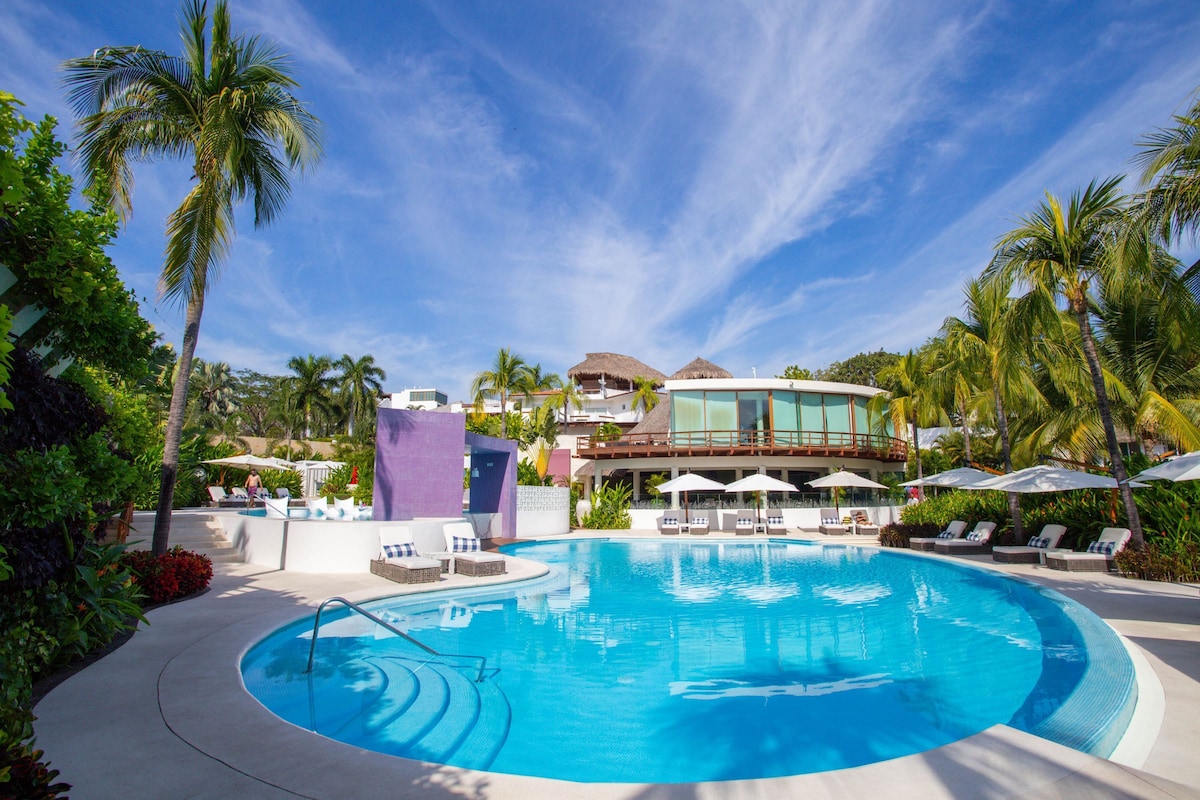 A resort with a large swimming pool, lounge chairs, and palm trees.