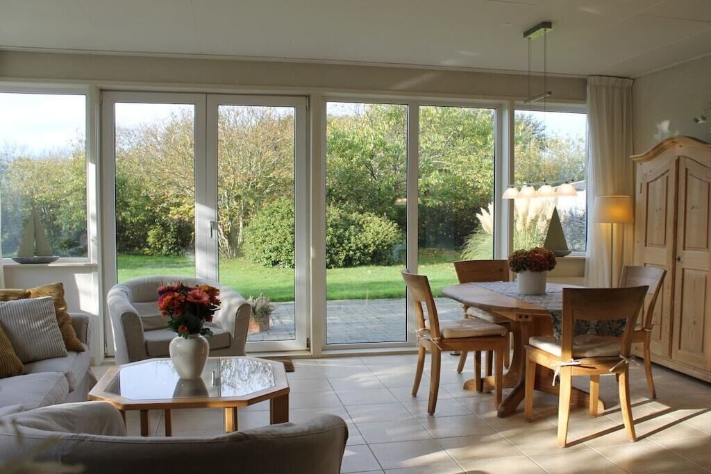 Een ruime woonkamer met een grote schuifpui naar een tuin, een eethoek met houten meubels en een witte bank met een bloevas.