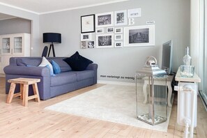 Uma sala de estar moderna com um sofá cinzento, um banquinho de madeira, uma mesa de vidro e uma parede decorada com quadros.