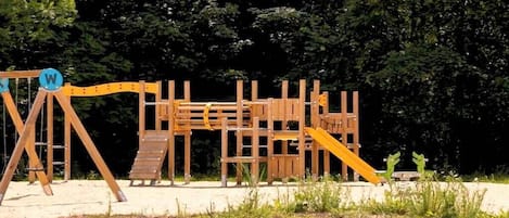 A wooden playground structure with a slide and climbing elements, surrounded by trees.