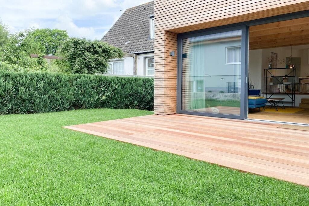 Una casa moderna con terraza de madera, puerta de vidrio que da a una habitación y césped cuidado.