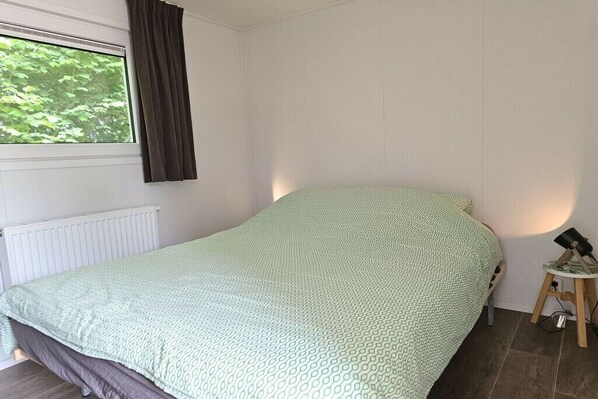 A single bed with a patterned bedspread in a room with white walls, a window with curtains, and a small bedside table with a lamp.