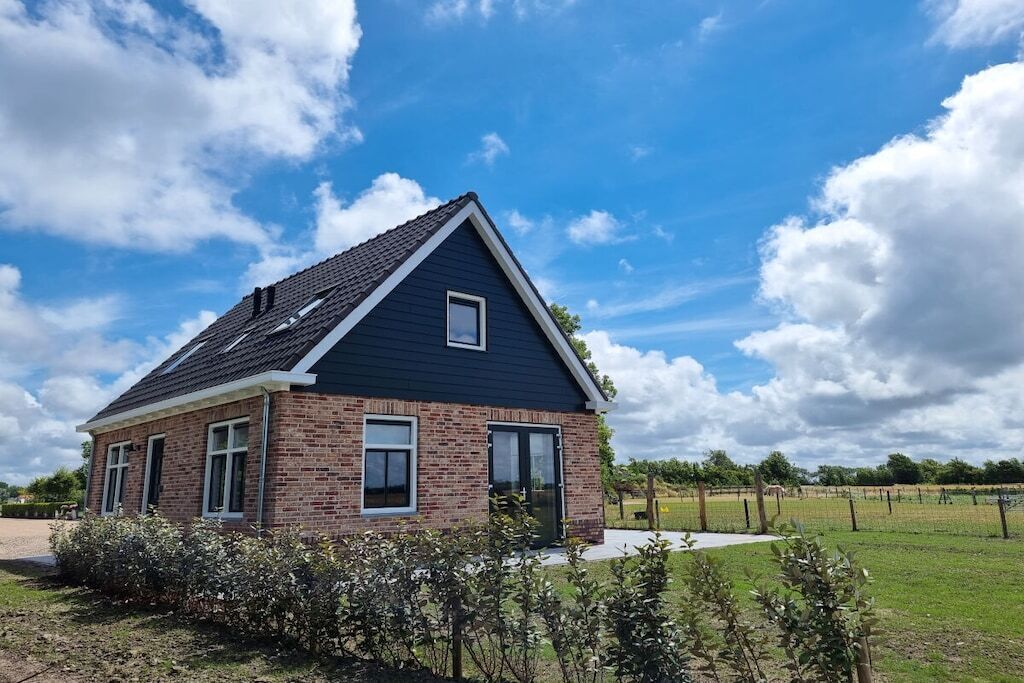 Une maison à deux étages, avec une façade en briques et un toit foncé, entourée d’une pelouse et d’un champ clôturé.