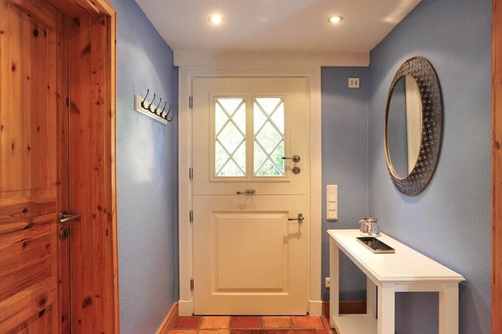 A hallway with a wooden door, a white door with a window, a white console table with a mirror, and a wall-mounted light fixture.