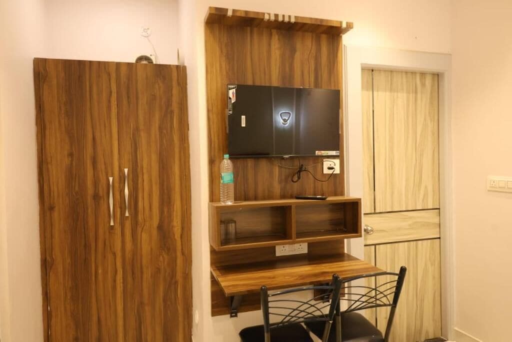 A room with a wooden wardrobe, a mounted television, and a small wooden table.
