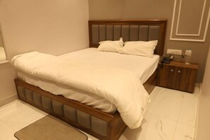 A neatly made bed with a wooden headboard and a small bedside table with a phone and a bottle. - HOTEL SHREE KARNI PALACE (Bulandshahr)