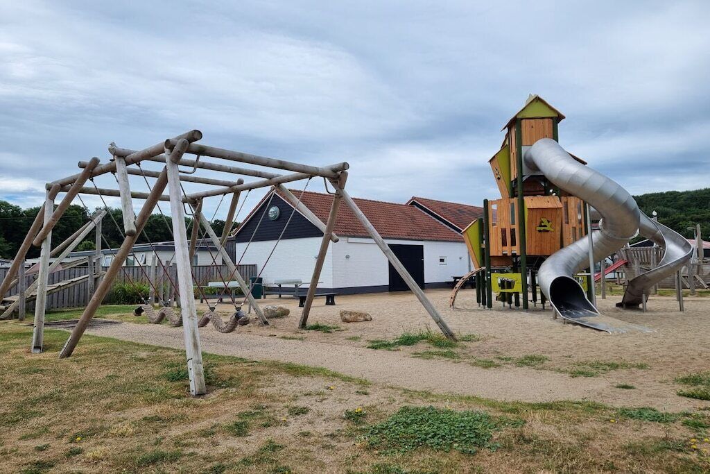 Une aire de jeux avec une balançoire en bois, un toboggan et un petit bâtiment doté d’un toit en pente.