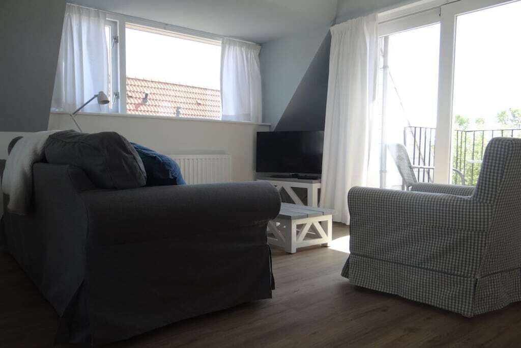 Ein Zimmer mit einer Couch, einem Sessel, einem Fernseher auf einem weißen Schrank und einem Balkon mit Geländer.