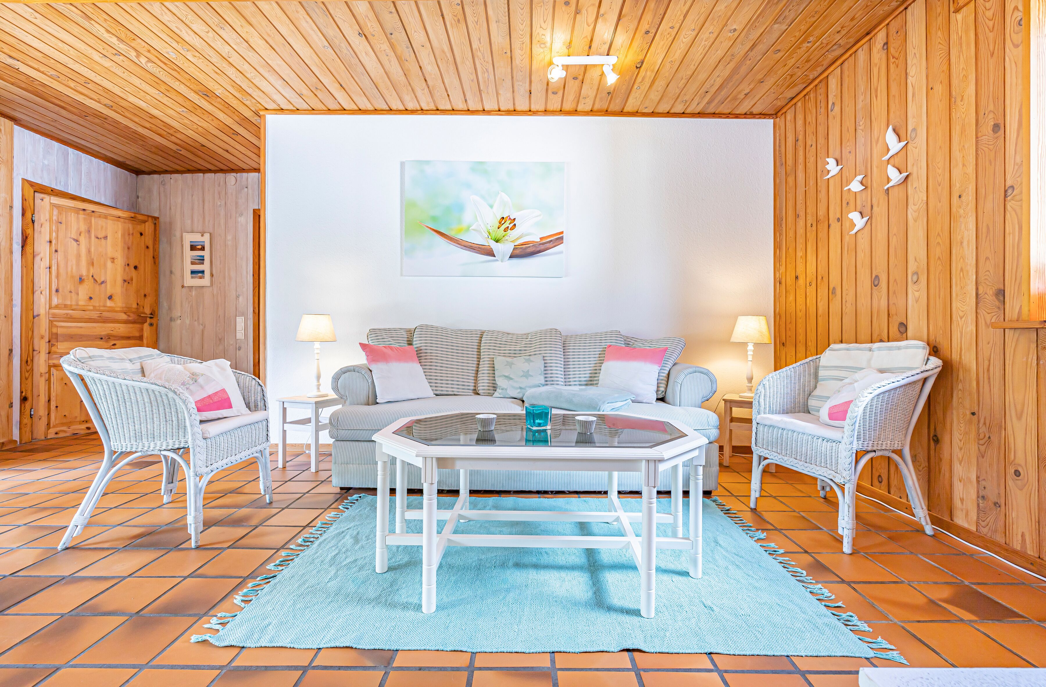 A cozy room with a wooden ceiling, a sofa set, two wicker chairs, a coffee table, and a blue rug.