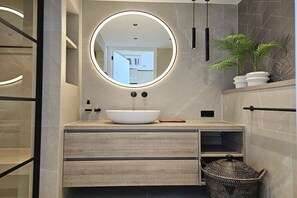 A modern bathroom with a round mirror, a white sink, wooden drawers, a potted plant, and a wicker basket. - Holiday apartment with 1 bedroom (Domburg)