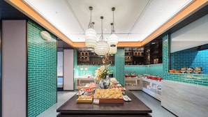 A modern interior with a central table displaying food items, a kitchen area with blue-tiled walls, and hanging pendant lights.