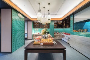 Un interior moderno con una mesa central que exhibe alimentos, una zona de cocina con paredes de azulejos azules y lámparas colgantes.