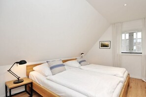 A bedroom with a wooden bed, white bedding, a nightstand with a lamp, and a window with curtains. - Holiday home with 1 bedroom (St.Peter-Ording)