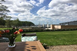 A wooden table with a vase of tulips, a bench, and a view of a marina with boats and a building in the background. - Marina-Fördeblick (Luck 2) (Glücksburg)