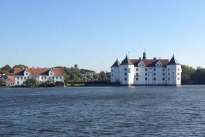 A large white building with multiple towers and a red roof, situated by a body of water. - Marina-Fördeblick (Luck 2) (Glücksburg)