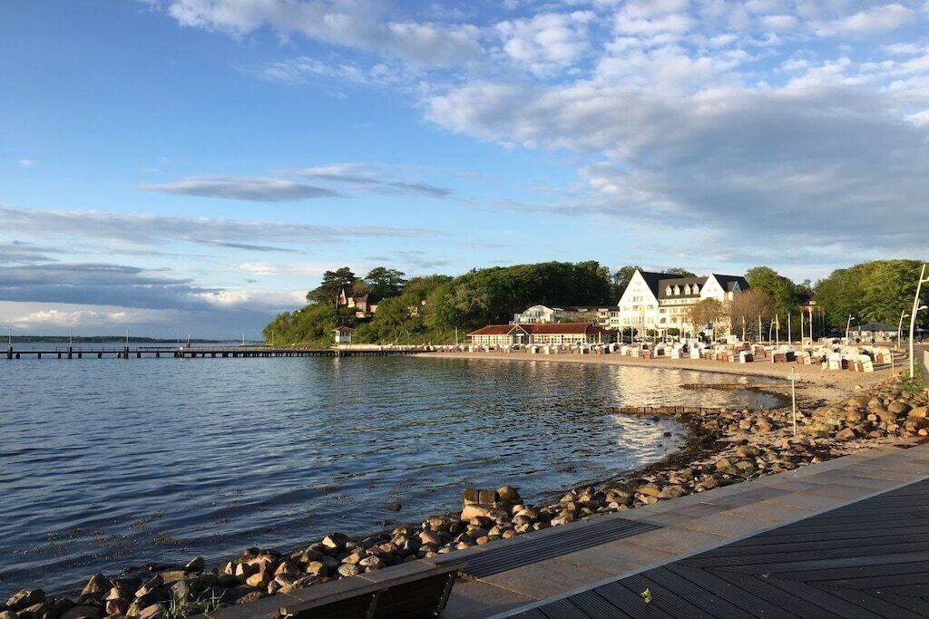 Ejendom ved vandet med træterrasse, stenet kystlinje og et hus med rødt tag.