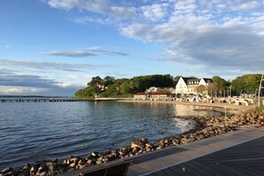 A waterfront property with a wooden deck, a rocky shoreline, and a building with a red roof. - Marina-Fördeblick (Luck 2) (Glücksburg)