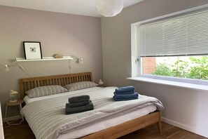 Une chambre avec un lit en bois, une fenêtre avec des stores, une table de chevet avec une lampe et une étagère murale avec un tableau.