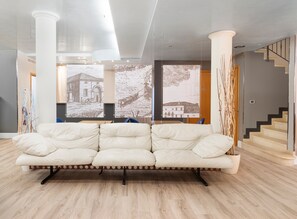 A modern living room with a white sofa, wooden flooring, and large windows with artwork.