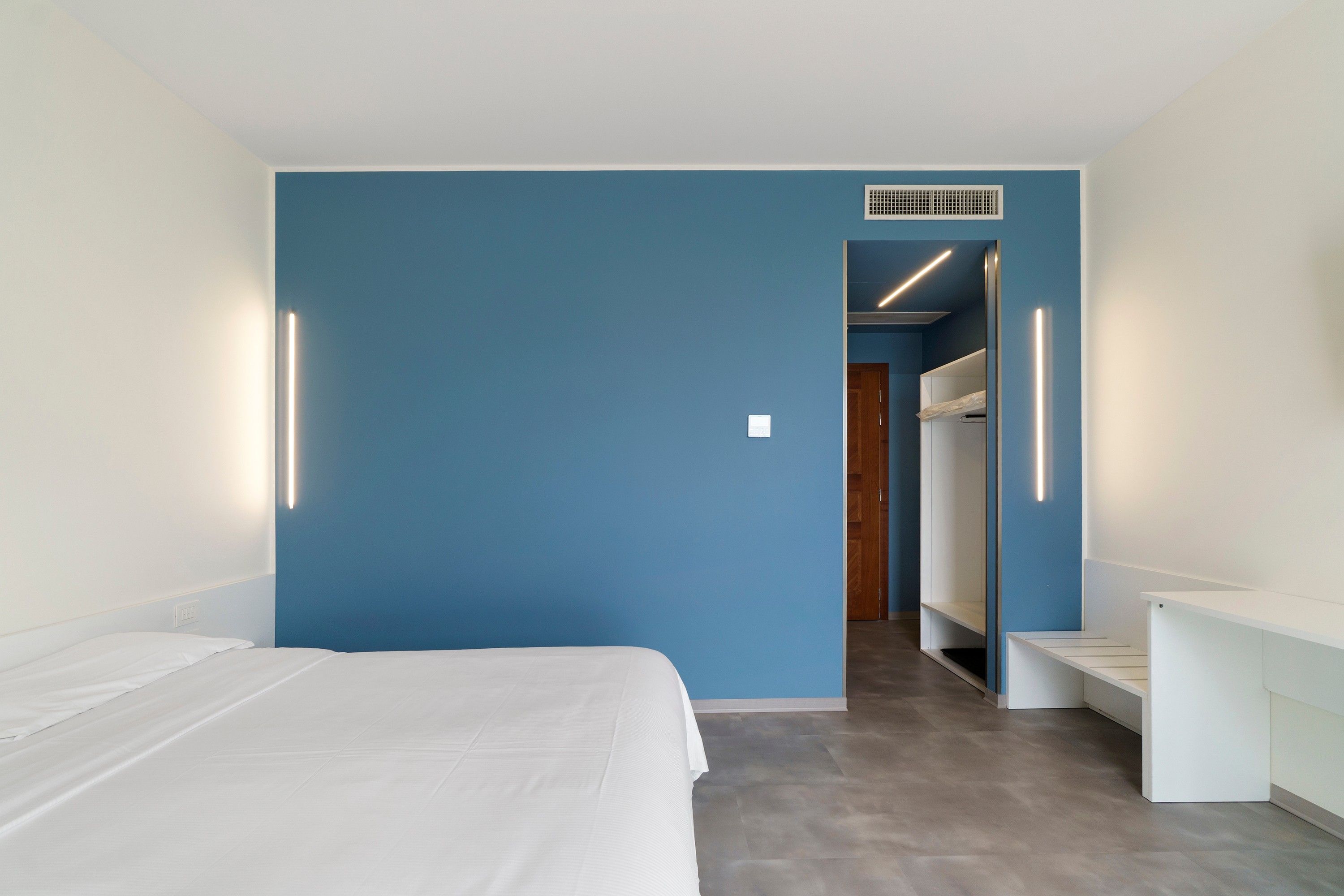 Une chambre d’hôtel moderne avec un grand lit, un mur d’accent bleu et un couloir menant à la salle de bain.