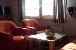 Una habitación con dos sillones rojos, una mesita con un cuenco, un televisor sobre un mueble y una ventana con cortinas.