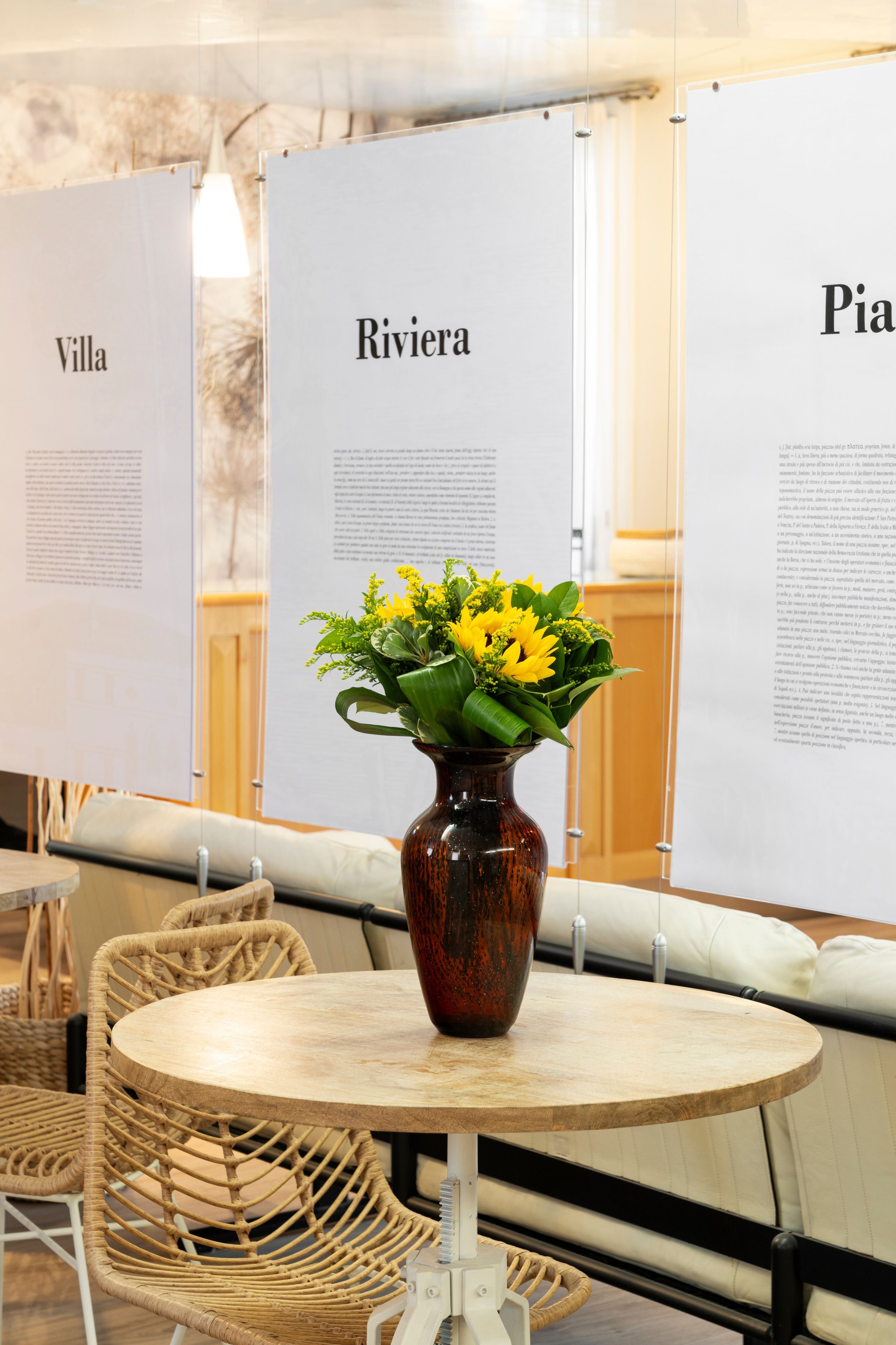 Ein runder Tisch mit einer Blumenvase voller gelber Blumen, ein Korbstuhl und eine Tafel mit Text.