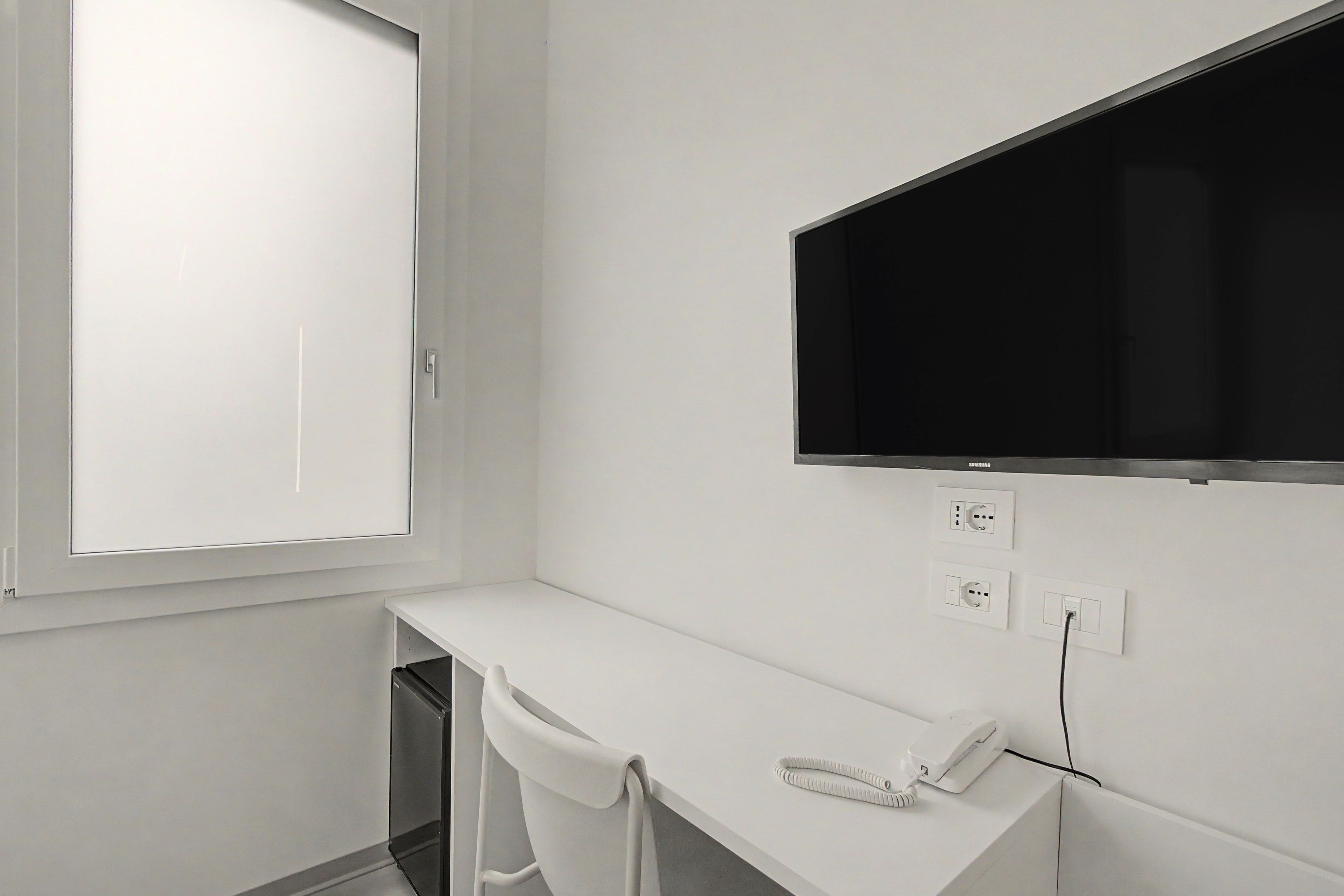 A white room with a window, a desk, a chair, a flat-screen TV mounted on the wall, and a telephone.
