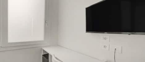 Habitación blanca con ventana, escritorio, silla, televisor de pantalla plana montado en la pared y teléfono.