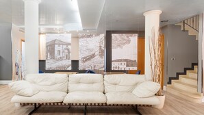 A modern living room with a white sofa, wooden flooring, and large windows with artwork.