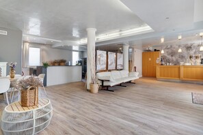 Un vestíbulo de hotel espacioso con suelo de madera, un sofá blanco, un mostrador de recepción y un gran ventanal.