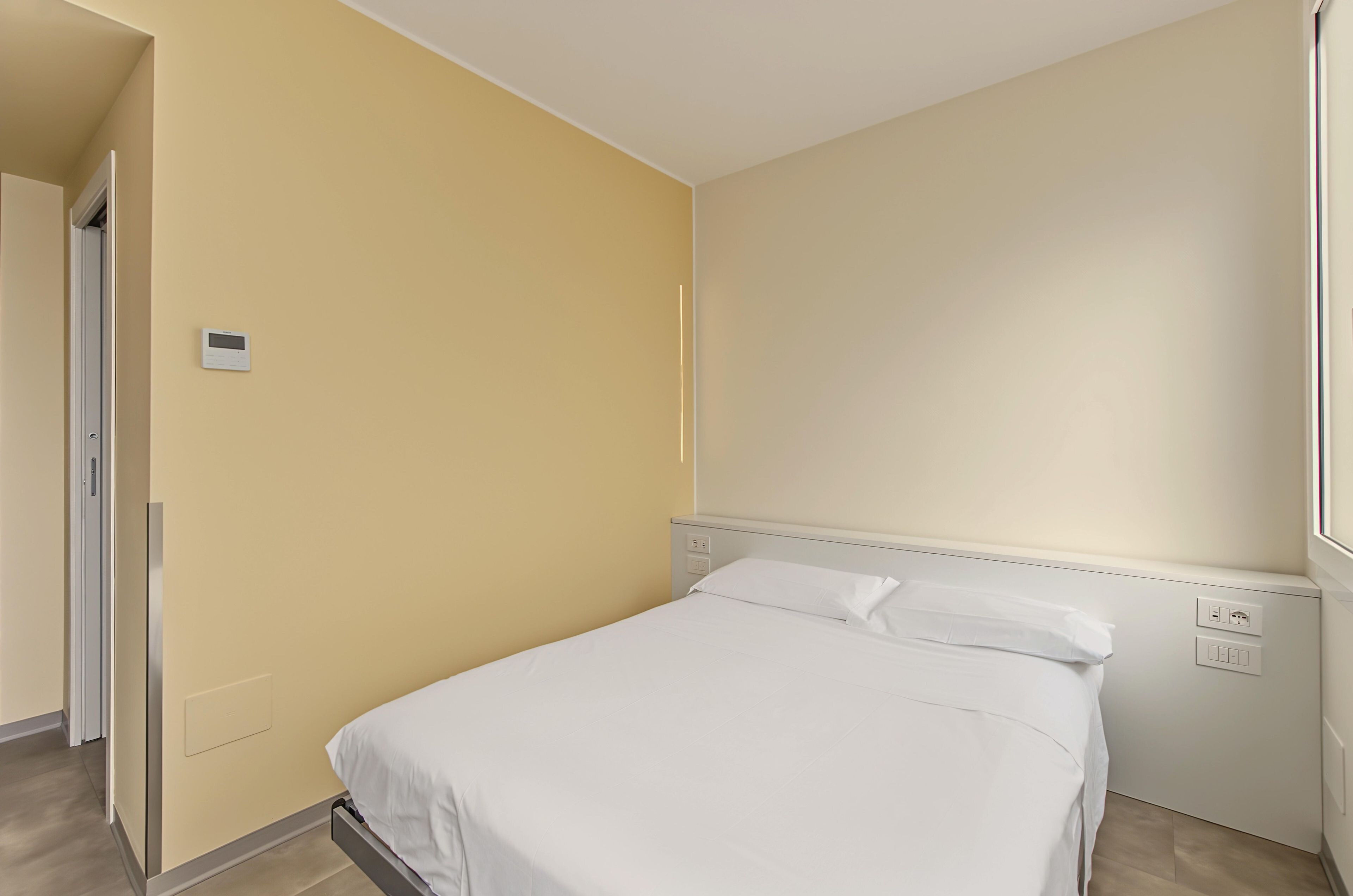 A bedroom with a bed, a wall-mounted thermostat, and a power outlet.