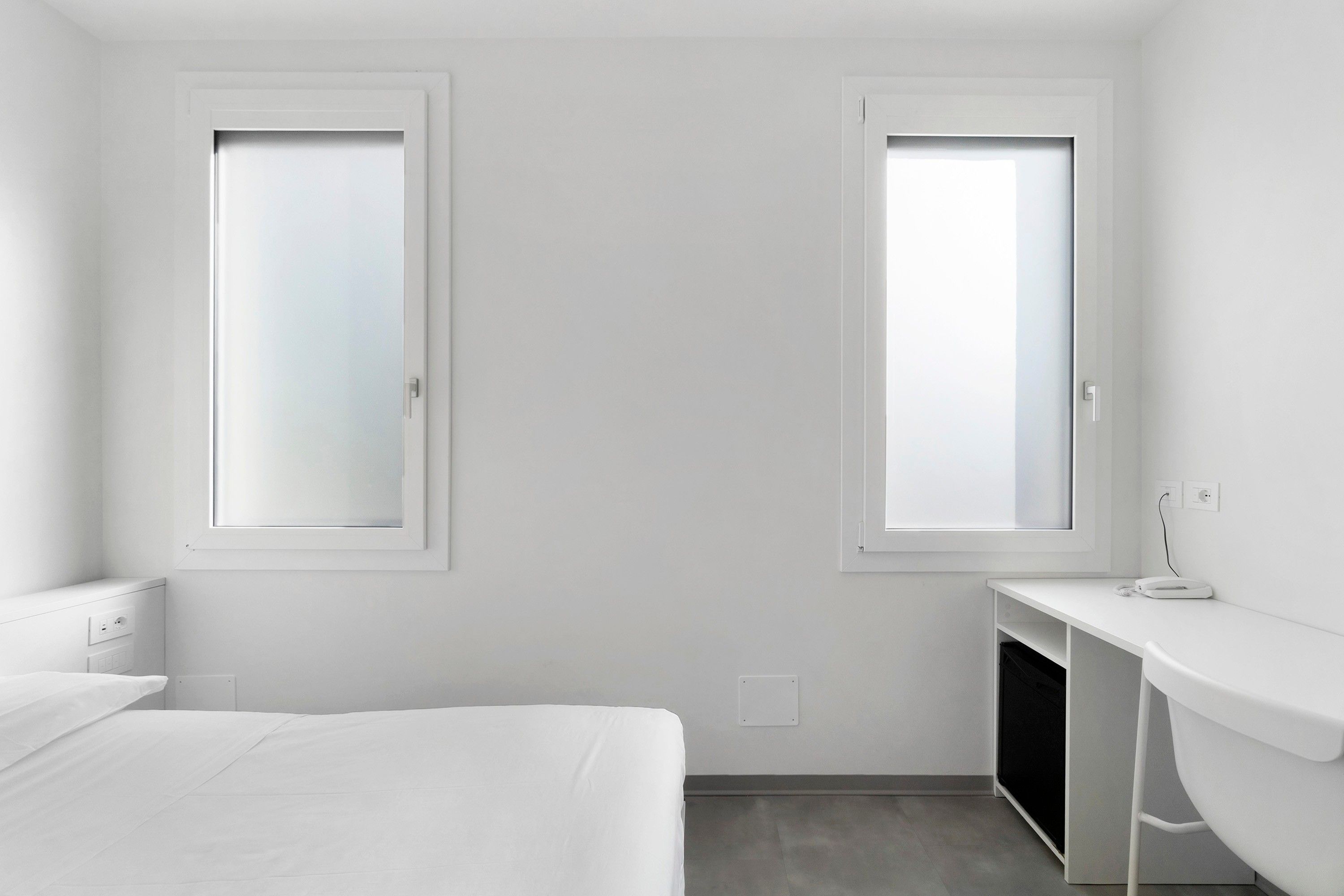 A minimalist hotel room with a bed, a desk, and two windows.
