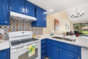 A modern kitchen with blue cabinets, a white stove, and a large window offering a view of the outdoors. - Monterey Country Club Condo | Golf Course & Lake Views, Gated Community, Heated Pools, Palm Desert (Palm Desert)