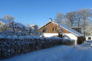 Ein Holzhaus mit geneigtem Dach, bedeckt mit Schnee, umgeben von schneebedeckten Bäumen und Sträuchern.
