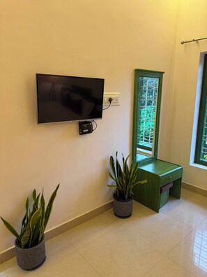A room with a flat-screen TV mounted on the wall, a green desk with a drawer, and two potted plants. - Hasu Stay (Madikeri)