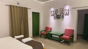 A hotel room with a bed, two chairs, a small table, a green door, and framed artwork on the wall. - Hasu Stay (coorg)
