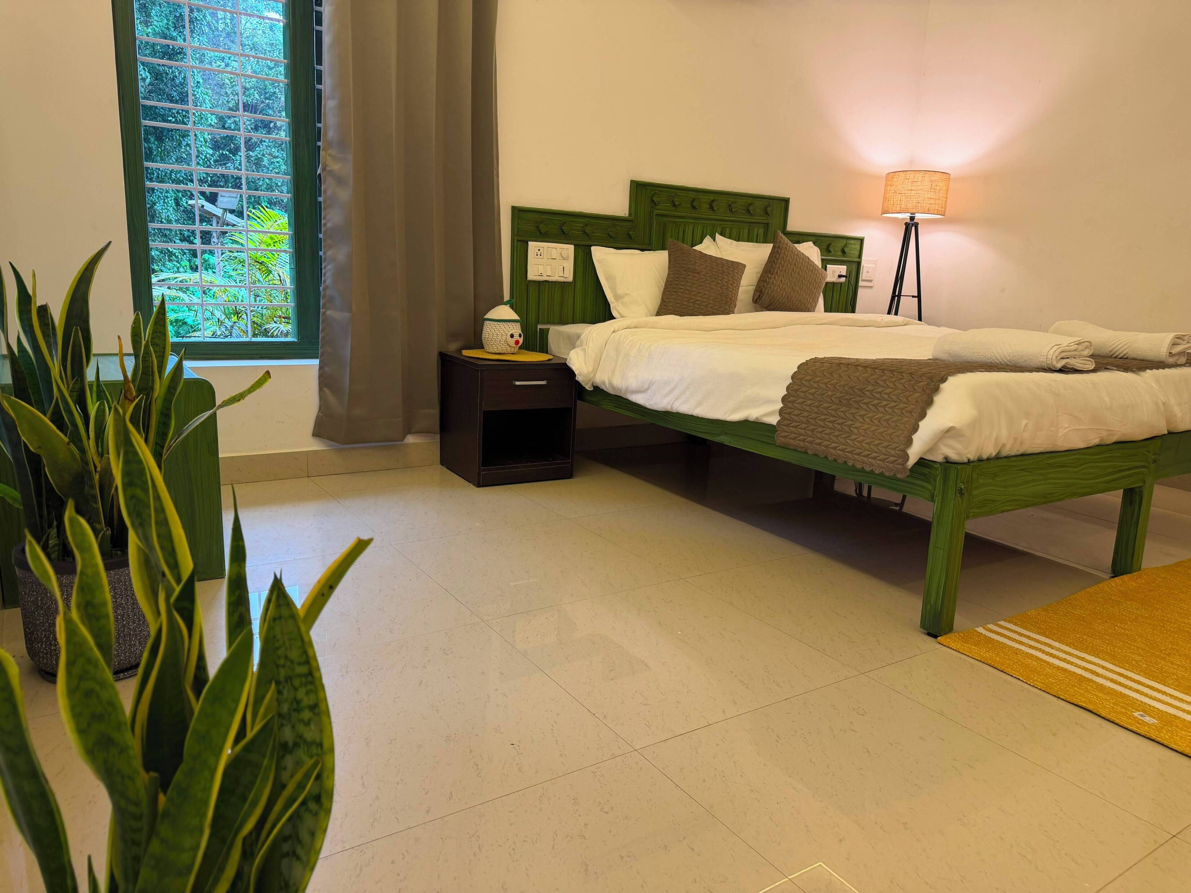 Una cama bien hecha con almohadas y una mesita de noche con lámpara, una ventana con cortinas y una planta en primer plano.