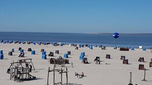 Uma praia com várias cadeiras e guarda-sóis azuis e castanhos, uma estrutura de madeira e um céu claro.