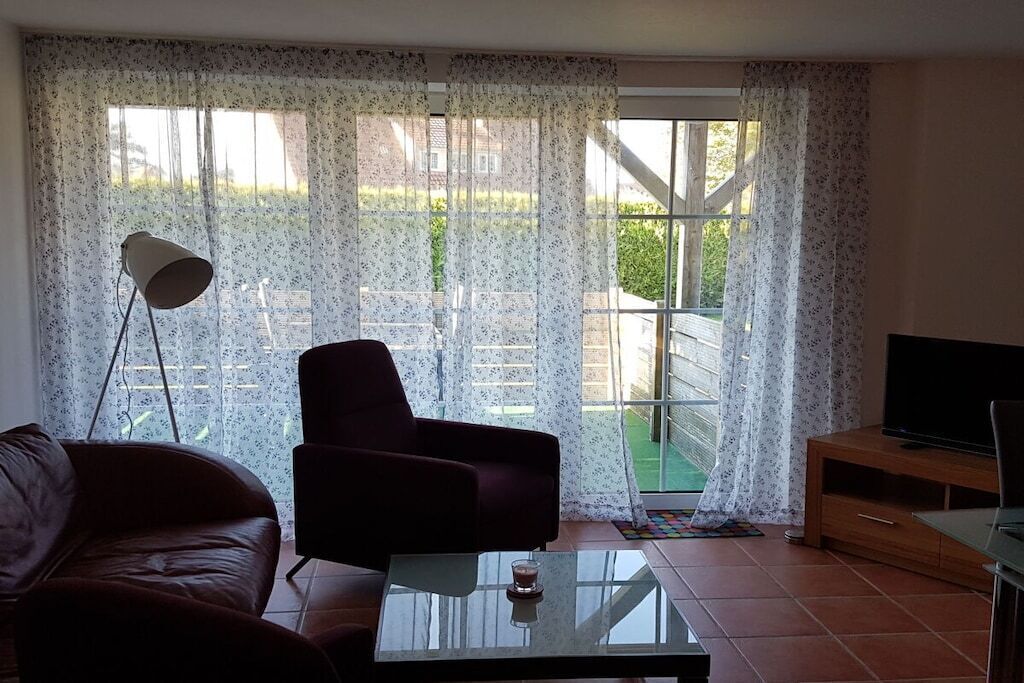 Ein Wohnzimmer mit Sofa, Sessel, TV-Schrank und einem großen Fenster mit Vorhängen.