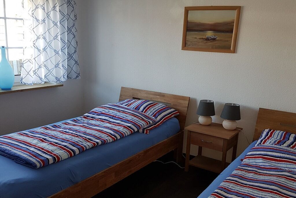 Quarto com duas camas de solteiro, uma mesinha de cabeceira com luminárias, uma janela com cortinas estampadas e um quadro na parede.