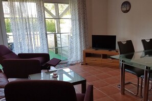Ein Wohnzimmer mit einer Couch, einem Glastisch, einem Flachbildfernseher und einem Esstisch mit Stühlen.
