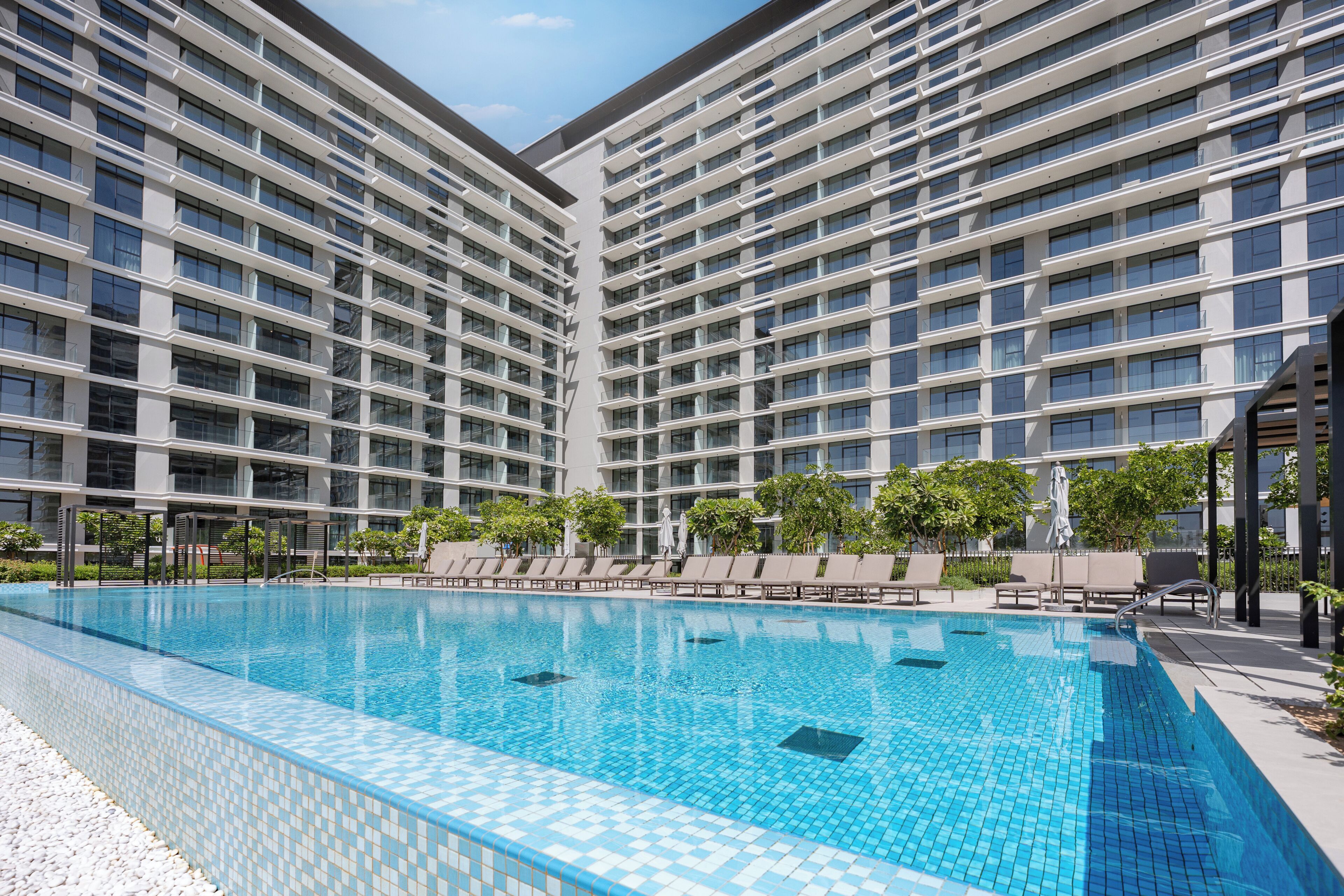 A modern hotel with a large pool area, multiple floors, and a clear sky.