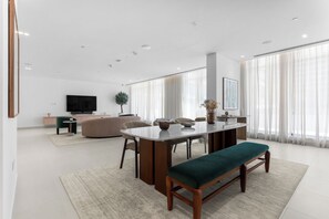 A modern living room with a dining table, seating, a flat-screen TV, and a large window with sheer curtains. - StayShort - Refined Classy 1 Bedroom in Dubai Hills - Sleeps 4! (Dubai Hills)