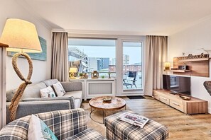 Una sala de estar moderna con sofá, reposapiés y mesa de centro, televisor y vistas de la ciudad a través de amplios ventanales.