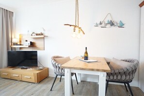 Una sala moderna con una mesa de comedor de madera, dos sillones tapizados, un televisor sobre un mueble de madera y un accesorio de luz fijado a la pared.