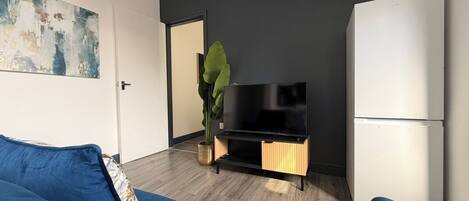 A modern living room with a blue sofa, a flat-screen TV on a wooden stand, a white refrigerator, and a large green plant.