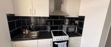 Una cocina compacta con armarios blancos, salpicadero de baldosas negras, estufa, microondas y pileta.