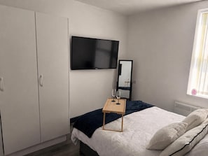 Un dormitorio con cama, televisión montada en la pared, armario y mesita de noche con vasos.