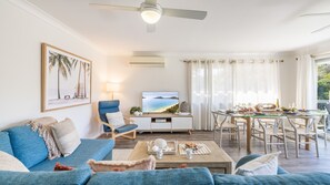A living room with a sofa, armchair, coffee table, TV, and dining area with chairs and a table. - Sunset Shores 1 Surf Air , 24 Marine Dr - air con , Wi-Fi, across to the beach (Fingal Bay)